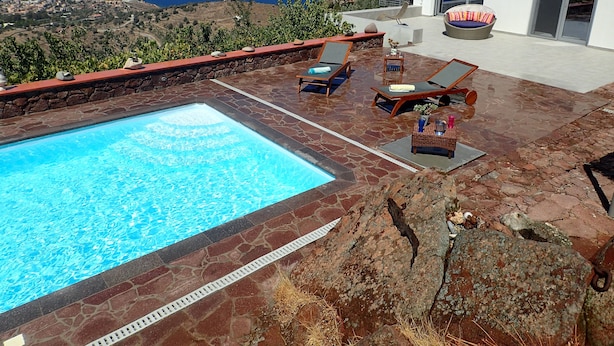 Área da piscina com vista para o mar, cadeiras de descanso e um edifício moderno.