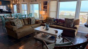 Ein gemütliches Wohnzimmer mit einem großen Fenster, das Blick auf das Meer bietet, einer Kristallleuchtre und einem Essbereich mit türkisfarbenen Stühlen.