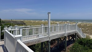 Eine Holzterrasse mit weißen Geländern blickt auf einen Strand mit Dünen und dem Meer.