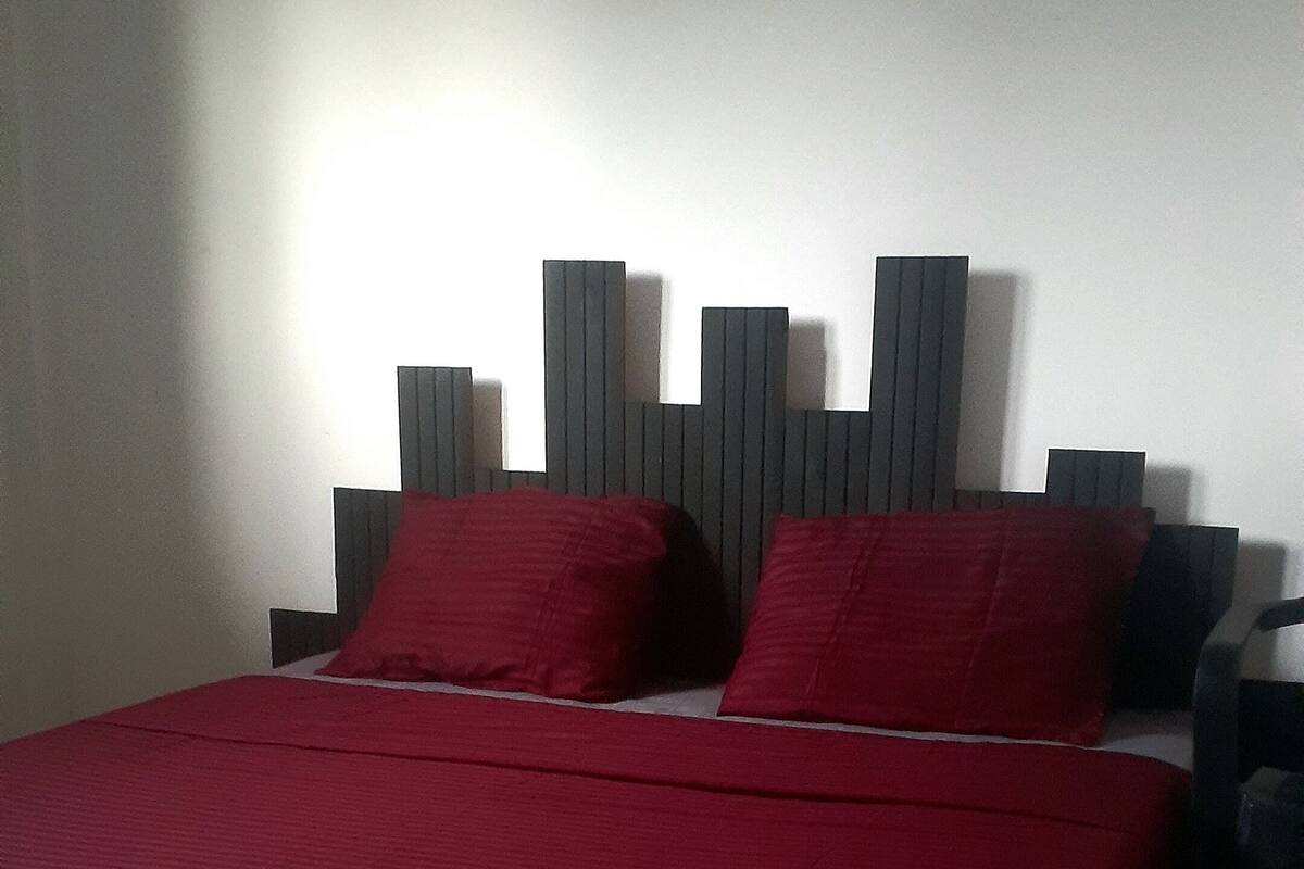 Ein Bett mit roter Bettdecke und zwei roten Kissen, ein hölzernes Kopfteil und ein Nachttisch mit einer Lampe.