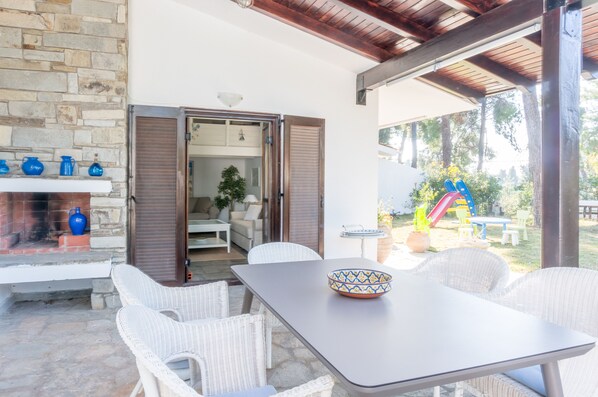 An outdoor patio area with wicker furniture, a stone wall, and a view of a playground. - Villa Aurora in Sani with Garden and Pool, by JJ Hospitality (Sane)