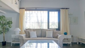Una sala de estar con un sofá beige, dos mesitas de noche blancas, una mesa de centro de vidrio, una planta en maceta y una ventana con cortinas.