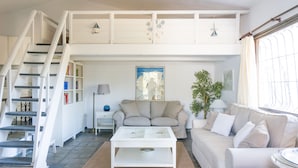 Un amplio salón con una escalera que conduce a un nivel superior, un sofá color beige, una mesa de centro blanca y una estantería.