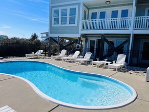 Une piscine avec des chaises longues, une terrasse en bois et un bâtiment à deux étages doté de balcons.