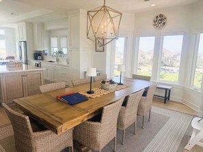 A spacious dining area with a wooden table and wicker chairs, a modern kitchen in the background, and large windows offering a view of the outdoors.