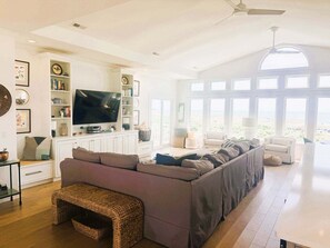 A spacious living room with a large flat-screen TV, built-in shelving, a long sofa, and a view through large windows.
