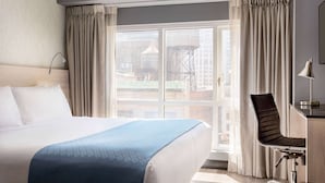 A hotel room with a large bed, a desk with a chair, and a view of buildings through the window.