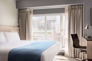 A hotel room with a large bed, a desk with a chair, and a view of buildings through the window.