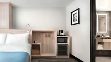 Une chambre d’hôtel avec un lit, une armoire en bois, un four à micro-ondes, un petit meuble, un tableau encadré au mur et une salle de bain visible à travers une porte ouverte.