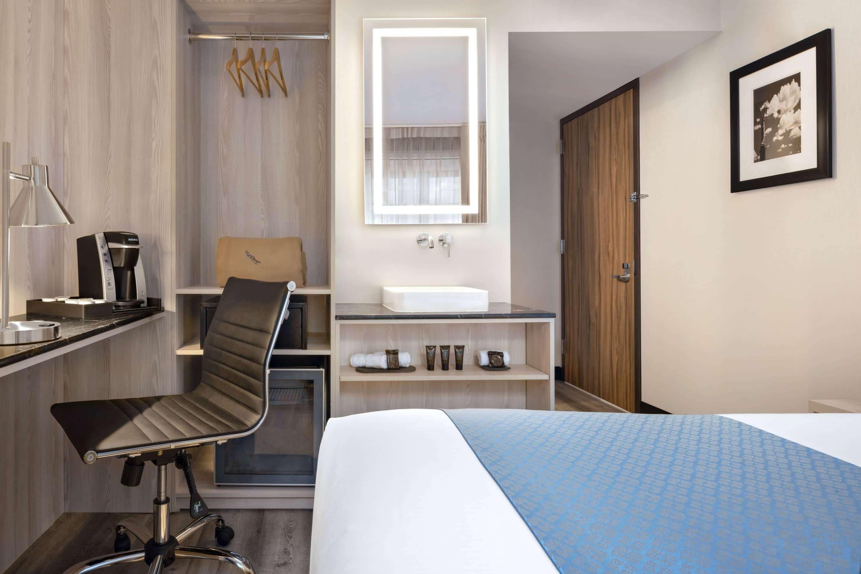 Habitación de hotel con cama, escritorio con silla, tocador con espejo, lavabo y armario.