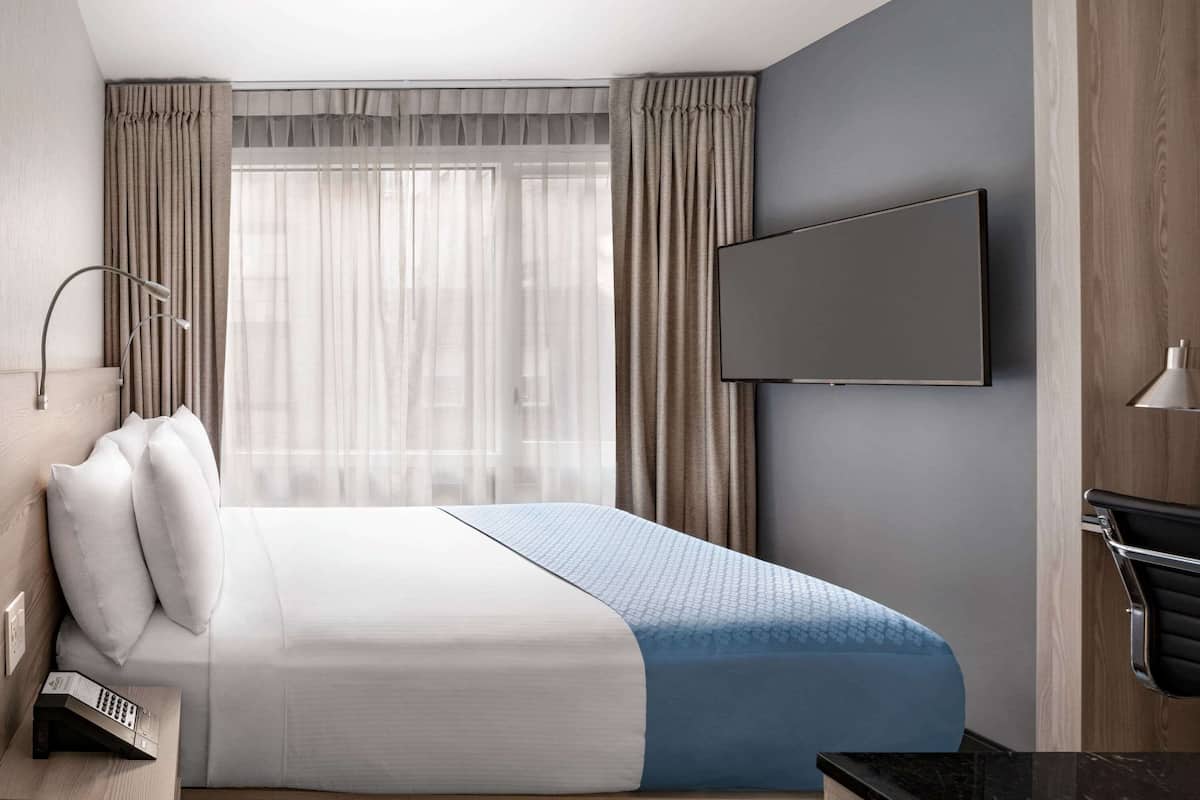 Une chambre d’hôtel moderne dotée d’un grand lit, d’un téléviseur à écran plat fixé au mur et d’une table de chevet avec une lampe et une télécommande.