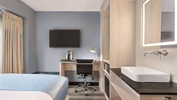 A hotel room with a bed, a desk with a chair, a flat-screen TV, a sink, and a mirror.
