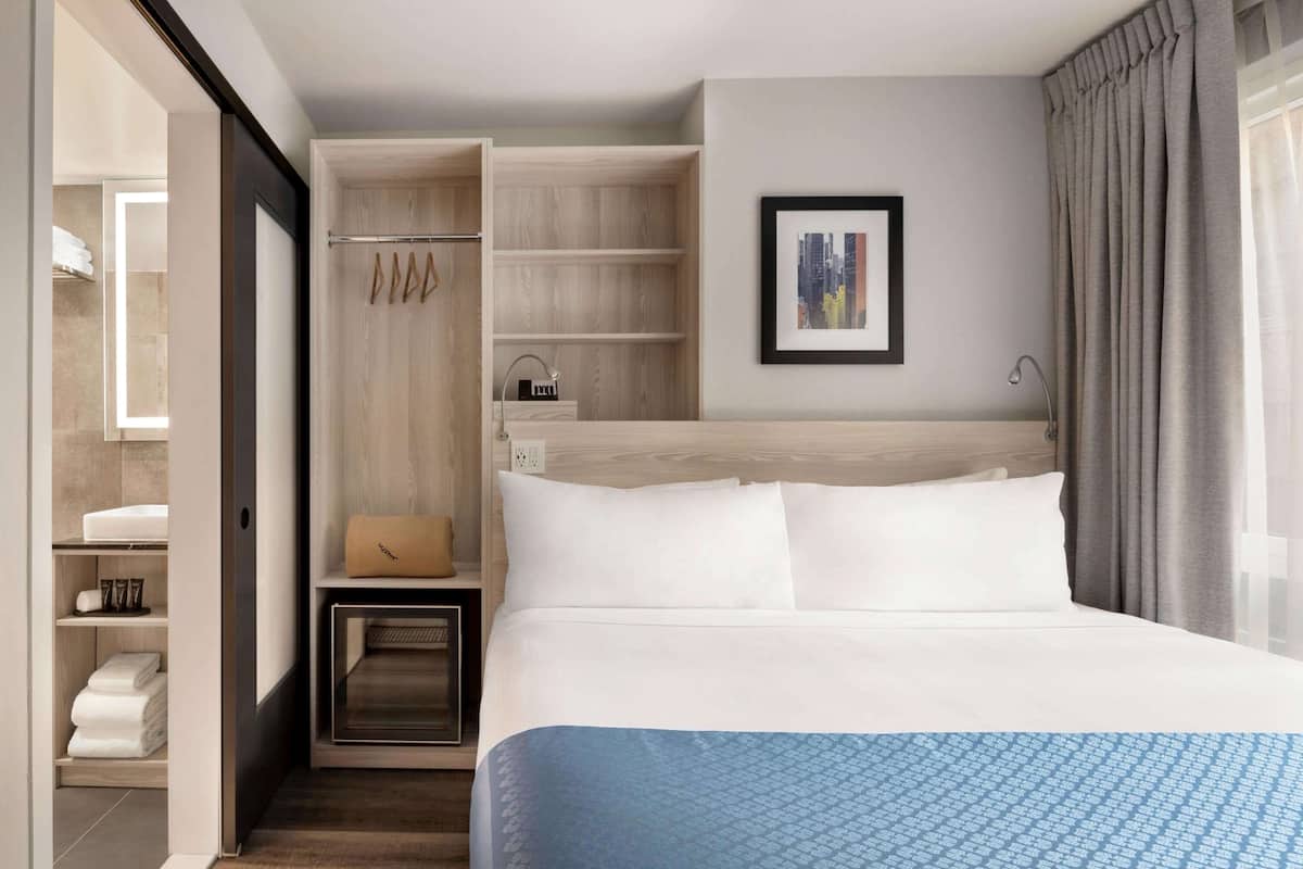 Une chambre d’hôtel avec un grand lit, un dressing, une salle de bain avec douche et un tableau représentant un paysage urbain accroché au mur.