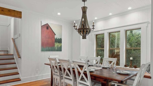 Un comedor con una mesa de madera, sillas blancas, una lámpara de araña y un cuadro de un granero.
