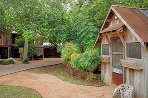 Een rustieke houten hut met een metalen dak, omringd door weelderige begroeiing en een grindpad.