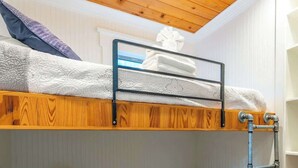 Una cama litera con techo de madera y barandillas de metal, un escritorio plegable y una pequeña estantería.