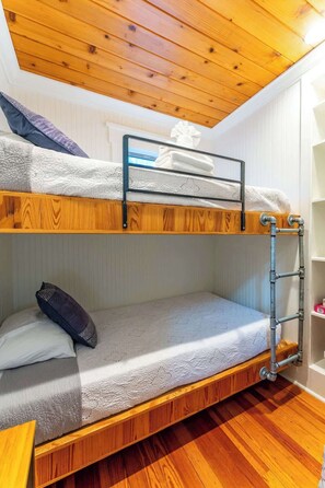 A bunk bed with a wooden ceiling and metal railings, a pull-out desk, and a small shelving unit.