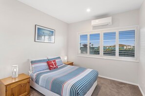 A bedroom with a bed, a nightstand, a lamp, a window with blinds, and an air conditioning unit. - Serenity 1 at Fingal Bay with Ocean Views (Fingal Bay)