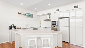 Une cuisine moderne avec des meubles blancs, un îlot central doté de tabourets, un four à micro-ondes, un four traditionnel et un réfrigérateur.