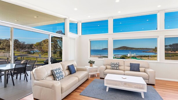 Un salon moderne doté d’une grande fenêtre donnant sur un paysage pittoresque, d’un canapé beige, d’une table basse et d’un coin repas avec des chaises.