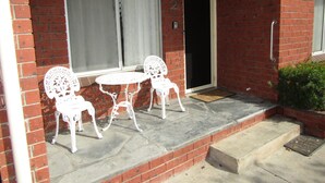 Un bâtiment en briques avec une porte blanche, une fenêtre ornée d’un rideau et un petit patio comprenant deux chaises blanches et une table.