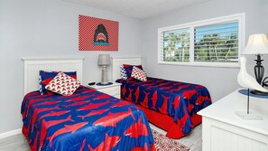 Une chambre avec deux lits, une table de chevet, une lampe, un tableau décoratif et une fenêtre donnant sur des palmiers.