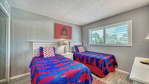 Una camera da letto con due letti, una finestra con persiane, una bocchetta di ventilazione a soffitto e un'opera d'arte appesa al muro.