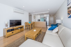 A modern living room with a flat-screen TV, wooden furniture, a sofa with blue cushions, and a kitchen in the background.