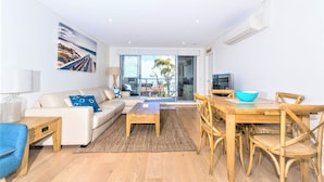 A modern living room with a sofa, coffee table, dining table, and chairs. There is a large window with a view of the outdoors. - Beautiful water views of Shoal Bay (Shoal Bay)
