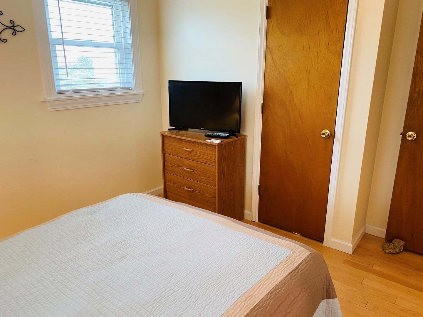A bedroom with a bed, a TV on a wooden dresser, a window with blinds, and a wooden door.