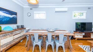 A dining area with a wooden table, grey chairs, a painting, a clock, and a TV. - Victor Parade Pet Friendly Shoal Bay Home (Shoal Bay)