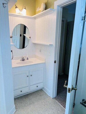 A bathroom with a white vanity, a round mirror, and a door leading to another room. - Experience coastal charm at Le Tournesol in Holden Beach, a spacious 4BR retreat! (Holden Beach)