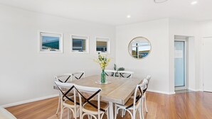 Ein Essbereich mit Holztisch, weißen Stühlen und einer Vase mit gelben Blumen. Drei Fenster bieten Blick auf das Meer, und an der Wand hängt ein runder Spiegel.