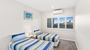 Een slaapkamer met twee bedden, een nachtkastje, een lamp, een lijst met een afbeelding, een raam met jaloezieën en een airconditioning.