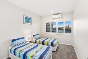 Quarto com duas camas, uma mesa de cabeceira, luminária, quadro emoldurado, janela com persianas e ar condicionado.