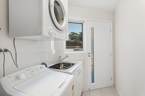 A compact laundry room with a front-loading washing machine, a built-in sink, and a window above the door. - Serenity 2 at Fingal Bay Waterfront Family Home (Fingal Bay)