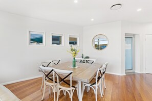 A dining area with a wooden table and white chairs, a vase with flowers, and three windows with views. - Serenity 2 at Fingal Bay Waterfront Family Home (Fingal Bay)