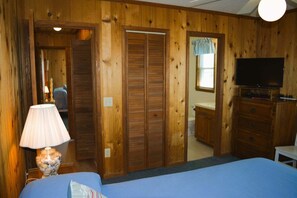 A room with wooden walls, a bed, a TV, a lamp, and a bathroom visible through an open door.
