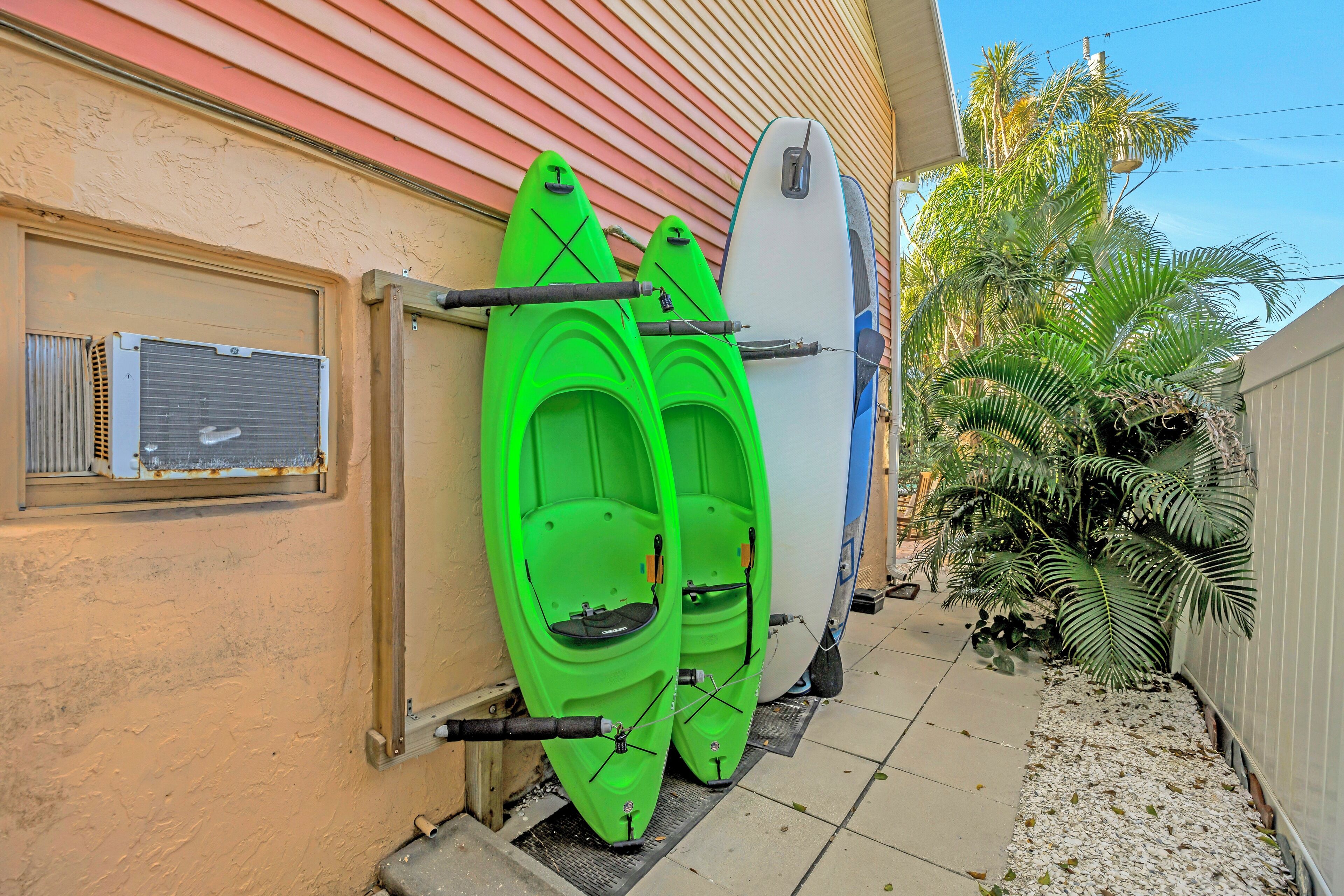 Deux kayaks verts et un kayak blanc sont garés contre un bâtiment, près duquel se trouvent une fenêtre et un palmier.