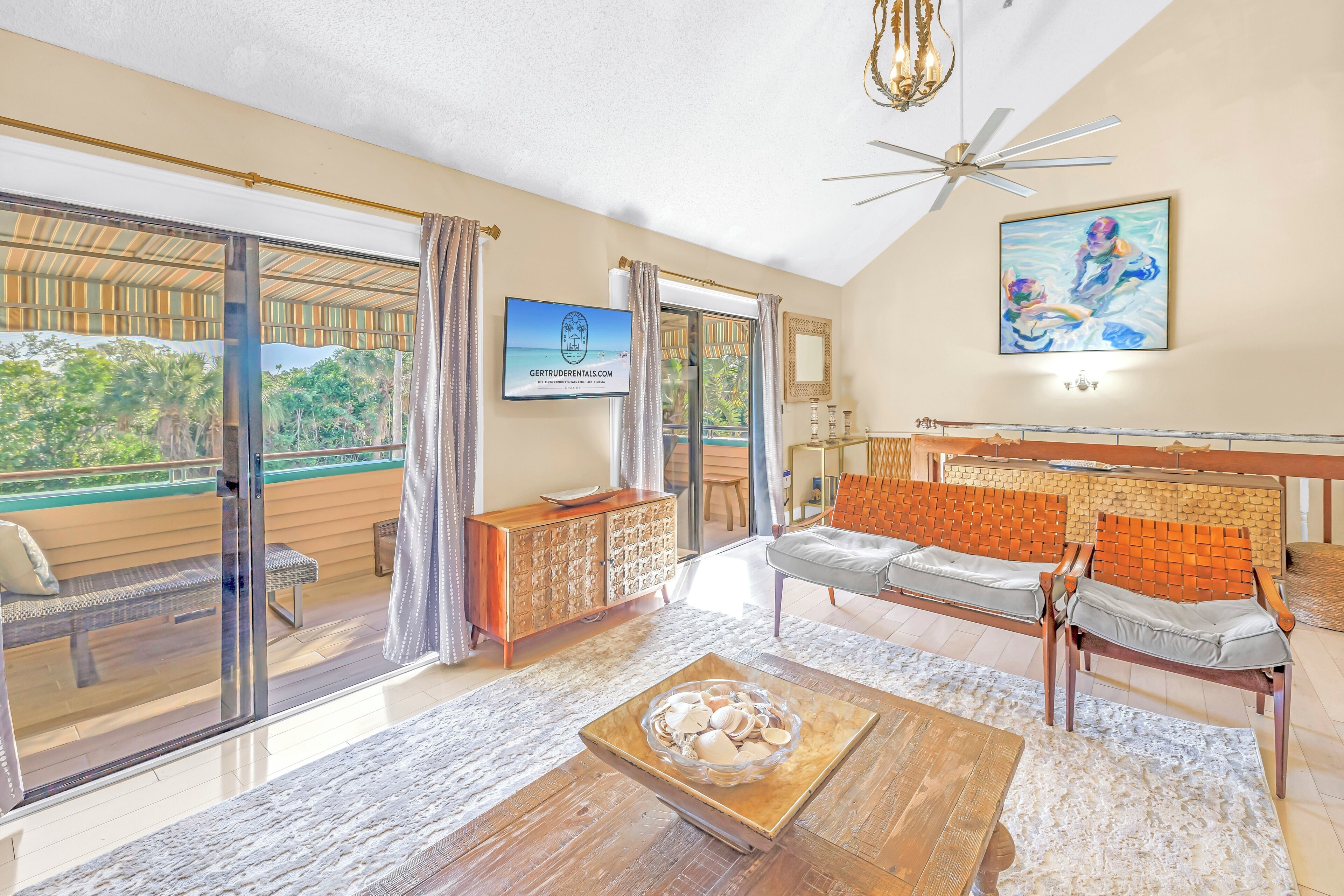 A spacious living area with a large sliding glass door leading to a balcony, a wooden coffee table, a sofa with cushions, and a television mounted on the wall.