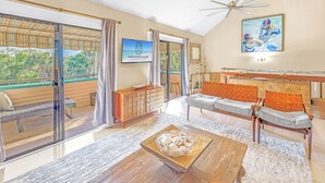 A spacious living area with a large sliding glass door leading to a balcony, a wooden coffee table, a sofa with cushions, and a television mounted on the wall. - Gertrude's Siesta | 4-min Walk to Beach & Village | Balcony & Tropical Patio! (Siesta Key)