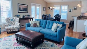 A living room with a blue sofa, a patterned armchair, a dark leather ottoman, and a colorful rug. - Stay at Wolfpack Waves, a spacious 4BR home in Holden Beach, perfect for 10 guests! (Holden Beach)