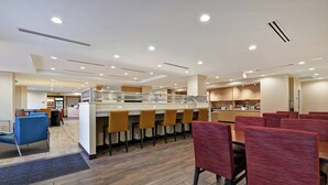 A modern hotel lobby with a central bar area, multiple seating options, and a visible staircase leading to an upper level.
