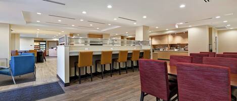 A modern hotel lobby with a central bar area, multiple seating options, and a visible staircase leading to an upper level.