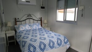 A bedroom with a bed, two bedside tables, a nightstand, an air conditioning unit, a window with blinds, and a wall-mounted light switch. - BLUE LINE UNIT 5 - Great location! close to all amenities! Off Peak Special! (Port Vincent)