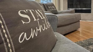 Ein Wohnzimmer mit Kamin, einer Couch mit einem Kissen, das "STAY awhile" sagt, und einem Schild über dem Kamin mit der Aufschrift "Relax LAKE TIME".
