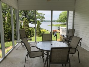 En overdækket terrasse med et rundt bord og fire stole, med udsigt over en sø med træer og en græsplæne.
