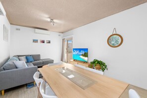 Ein modernes Wohnzimmer mit einem grauen Sofa, einem Holzesstisch und einem Fernseher, der eine Strandansicht zeigt.