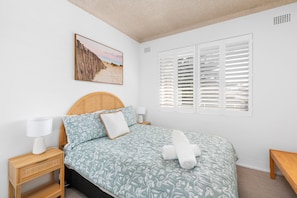 A bedroom with a bed, a bedside table, a lamp, a window with shutters, and a wall-mounted picture.