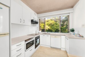 A modern kitchen with white cabinetry, a built-in oven, and a window with a view of trees. - Del Rae 15 Shoal Bay Apartment (Shoal Bay)
